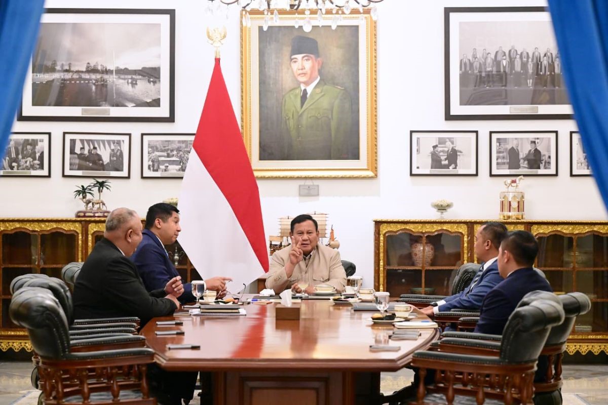 Group of officials sit around a polished wooden conference table in a formal meeting with the indonesian flag behind them and framed portraits on the wall behind the center speaker